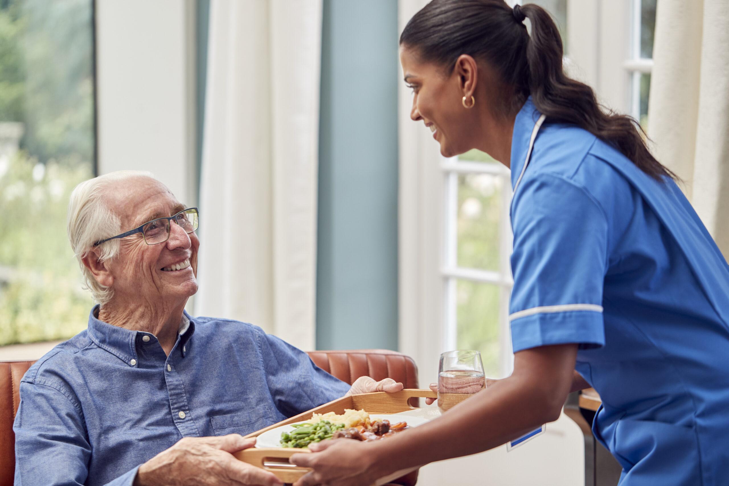 Female Care Worker In Uniform Bringing Meal On Tray To Senior Man Sitting In Lounge At Home